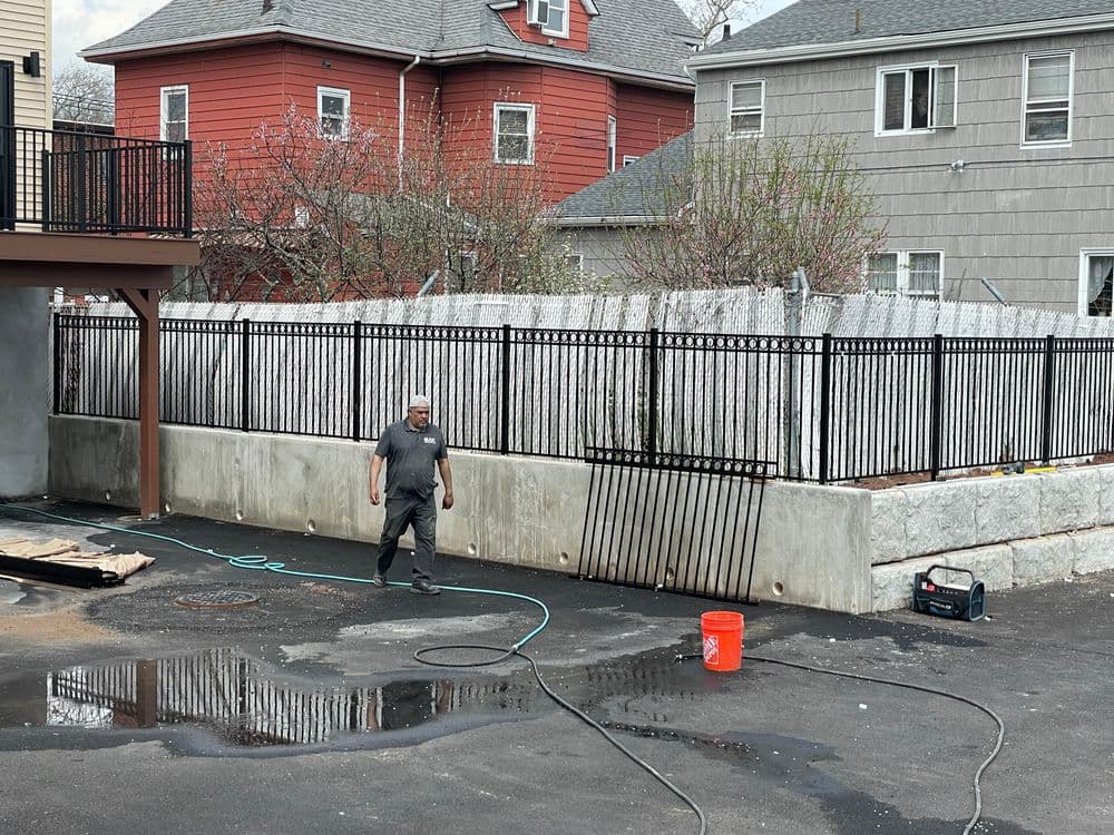 Worker cleaning outdoor area with water hose beside a fenced concrete wall and nearby houses.