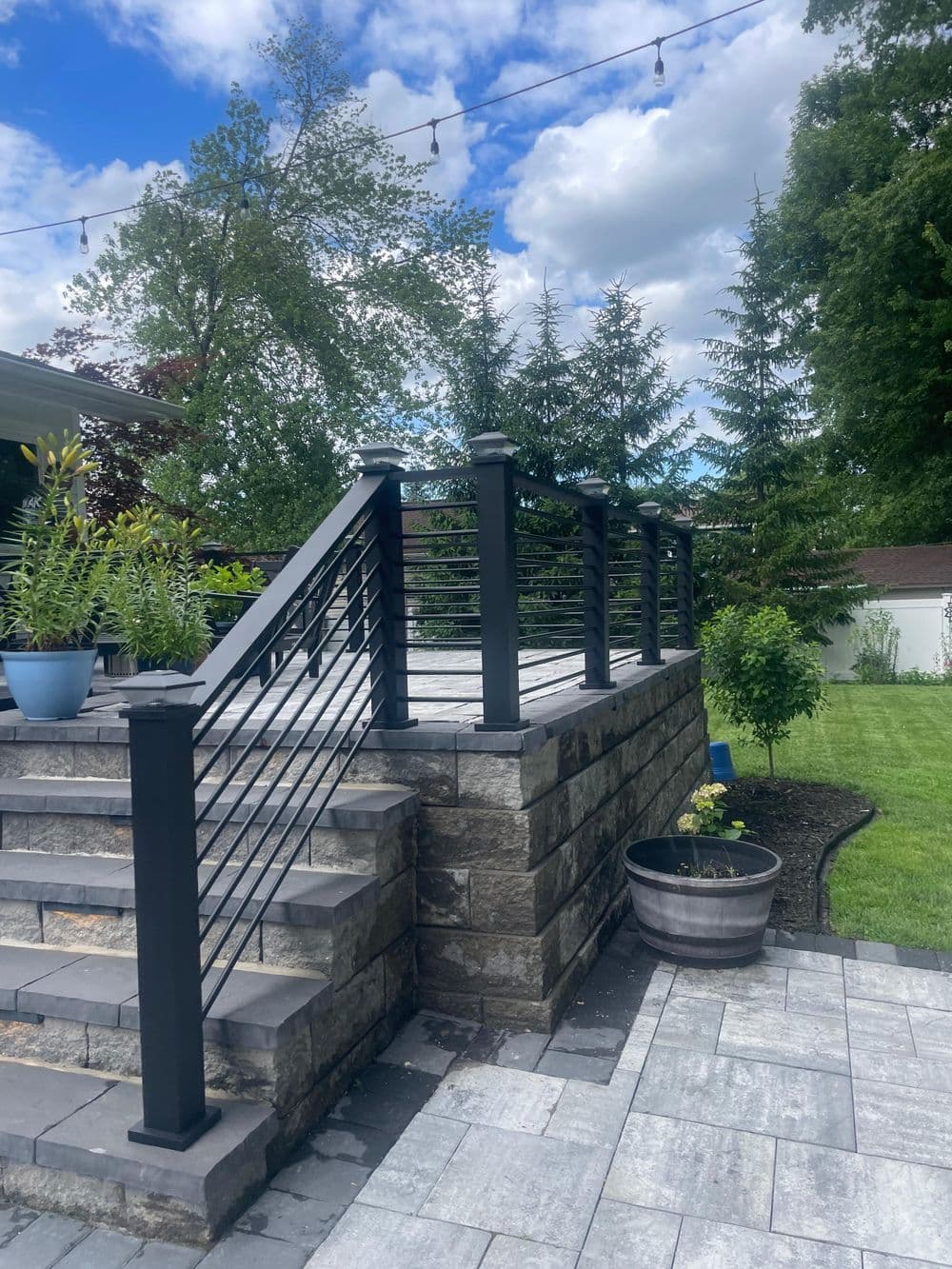 Modern outdoor staircase with black railing, stone walls, and lush greenery under a blue sky.