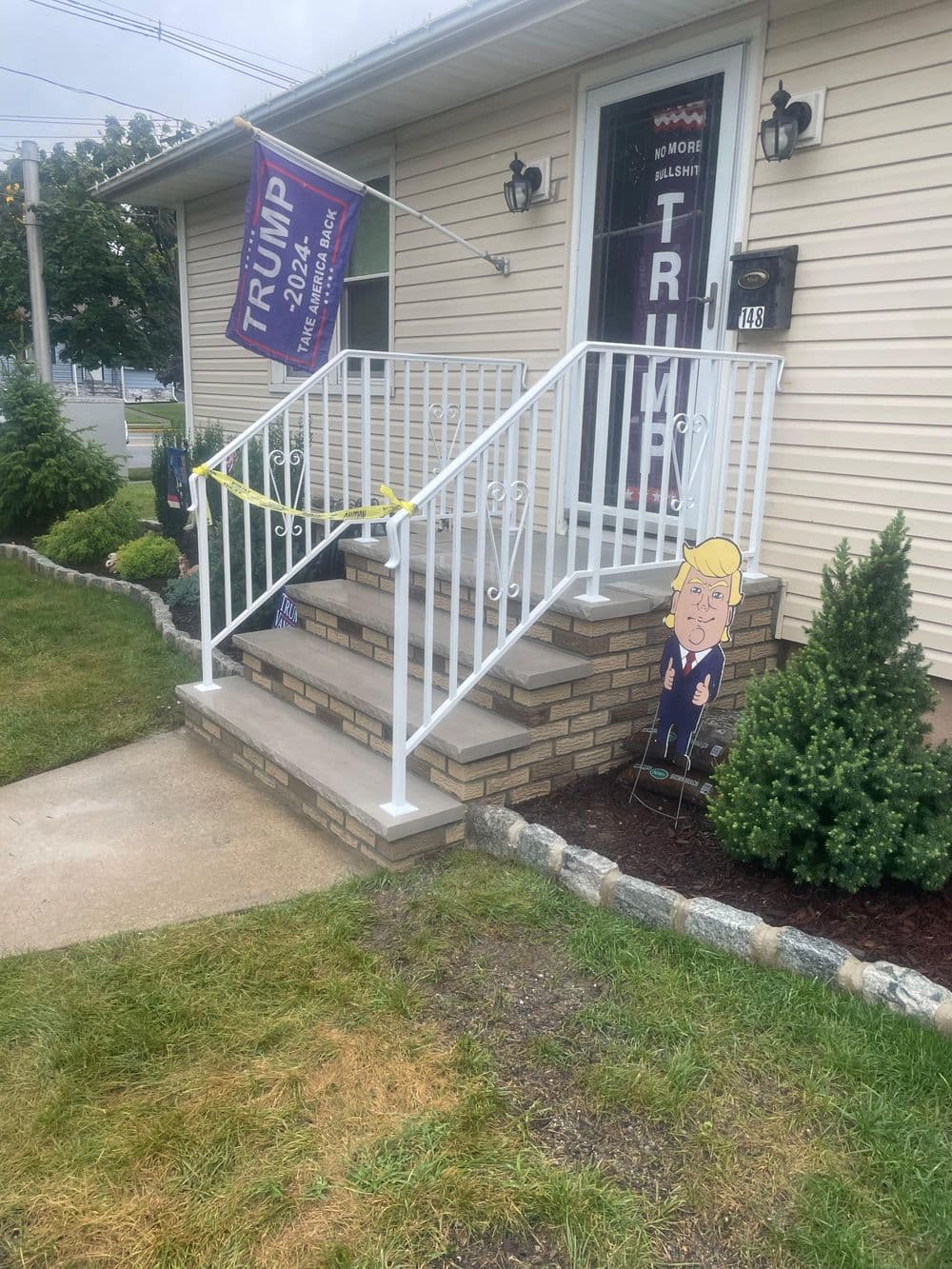 Trump 2024 campaign flag and cardboard cutout at a home entrance with landscaped yard.