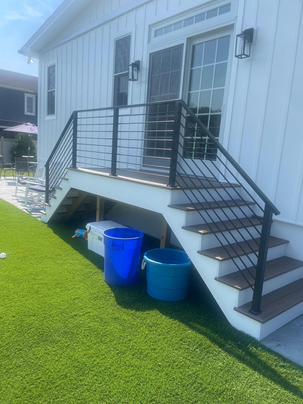 Modern deck with black railing, steps, and blue bins on a green lawn. Bright sunny day.
