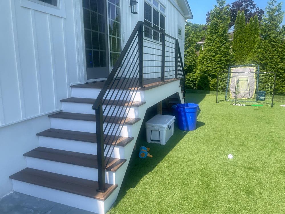 Modern outdoor stairs with a railing, leading to a white house, surrounded by green grass.