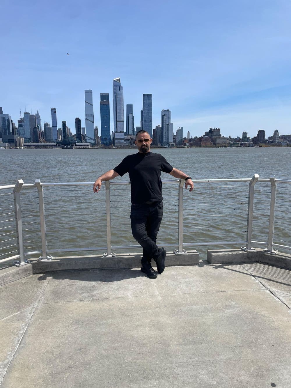 Man in black clothing poses on a waterfront with a city skyline in the background.