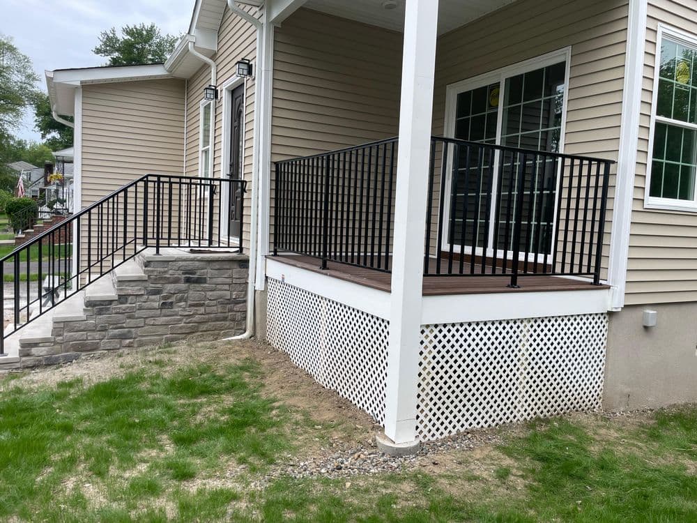 New home exterior featuring a black railing, wooden deck, and stone steps near green lawn.