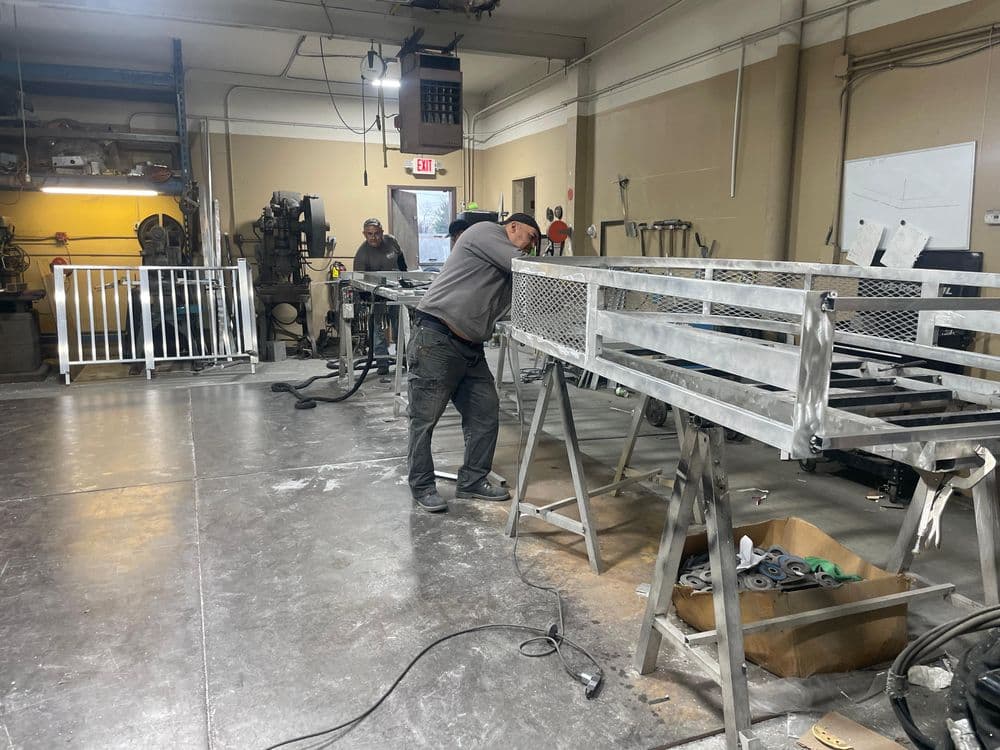 Worker assembling metal frame in a workshop with tools and equipment visible.