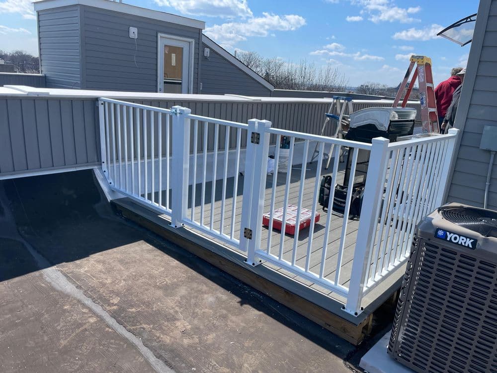 Rooftop deck with white railing, grill, tools, and ladder against a blue sky.