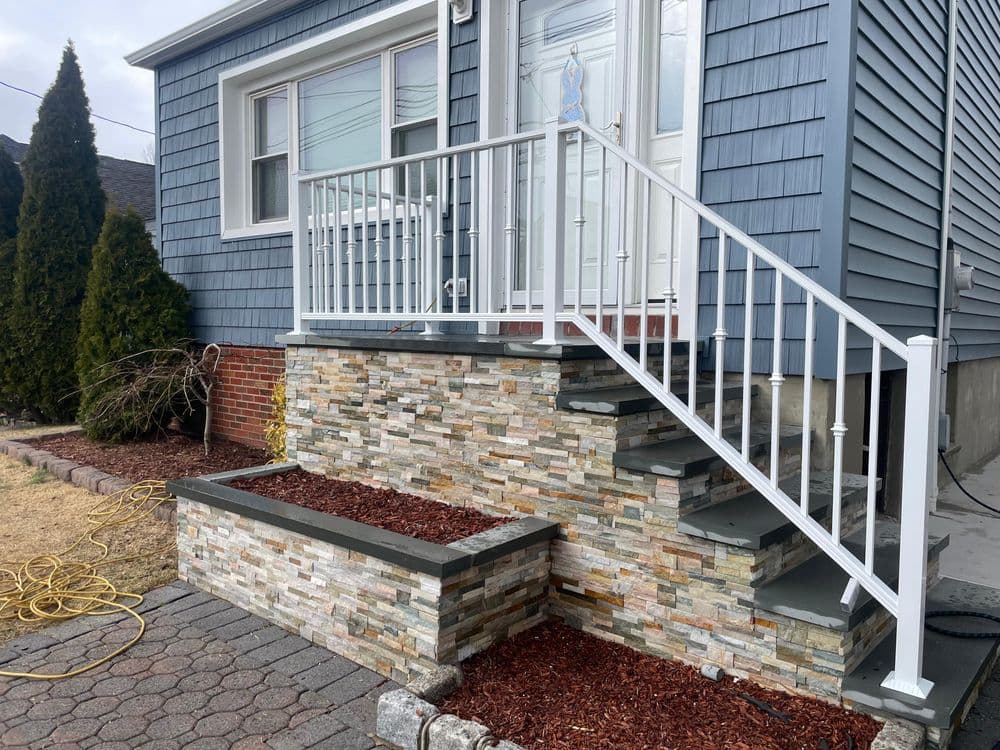 Entrance with stone steps, white railing, and landscaped flower beds at a blue house.