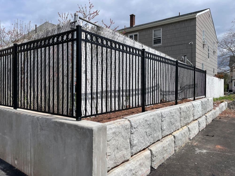 Black metal fence surrounding a stone wall garden area next to residential buildings.