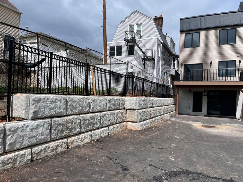 Retaining wall with fence near residential buildings and parking area on a cloudy day.