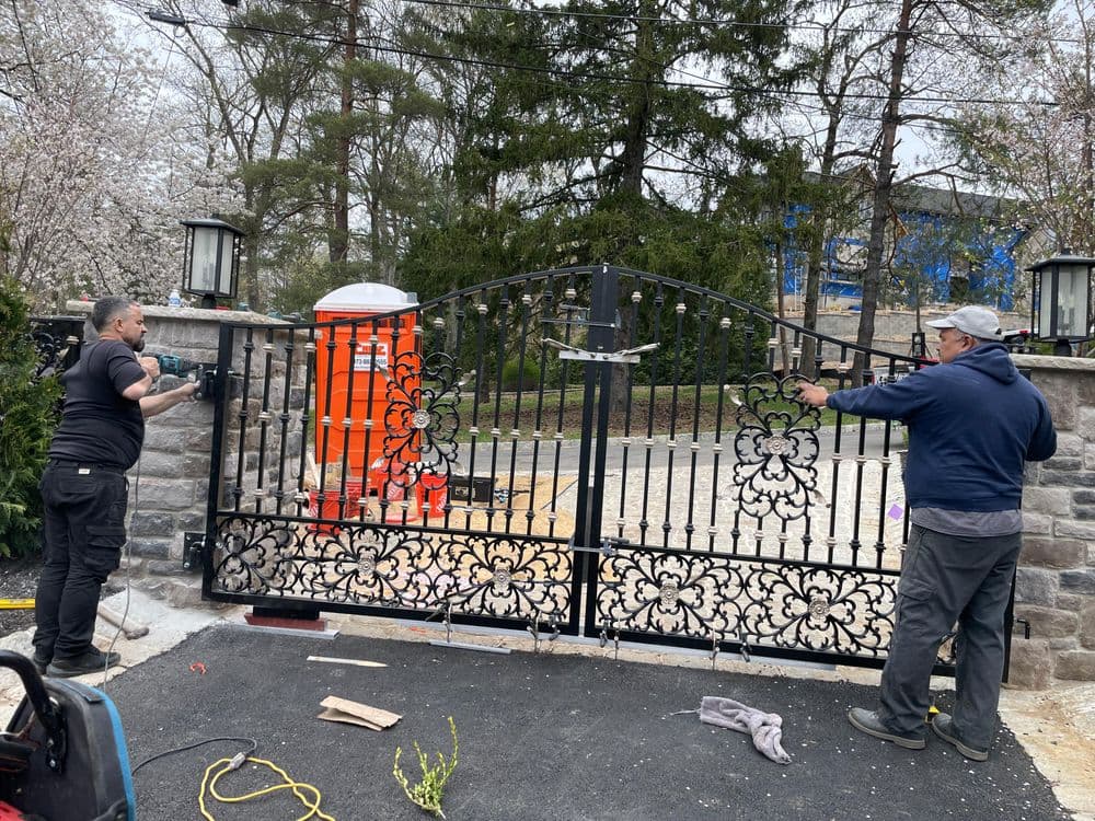 Two men installing an intricate metal gate at a property entrance, surrounded by trees.