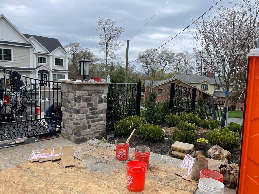 Stone gate entrance with decorative ironwork, residential landscape, and construction materials.