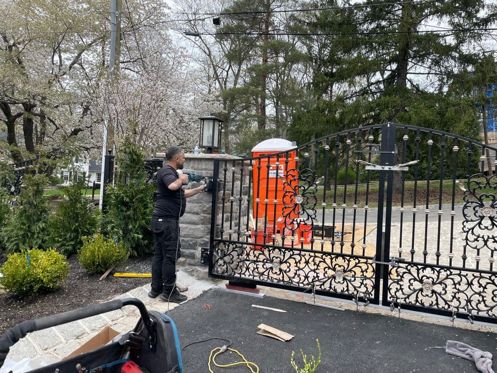 Worker installing a decorative metal gate at a residential entrance with blooming trees nearby.