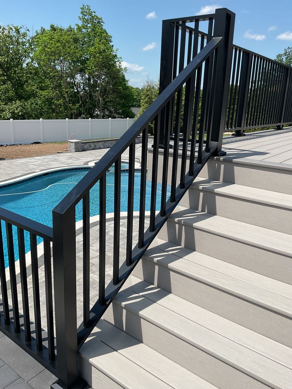 Modern black railing on stairs leading to a pool area, surrounded by greenery and blue sky.