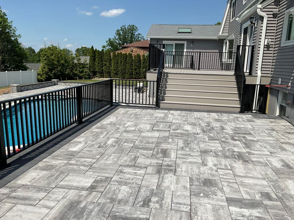 Modern backyard patio with gray stone tiles, black railing, and swimming pool in the background.
