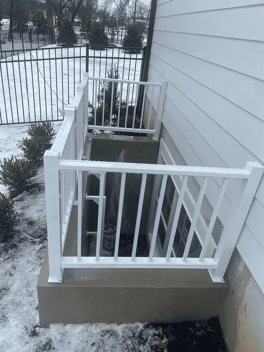 White railing on a basement entrance, surrounded by snow and greenery.