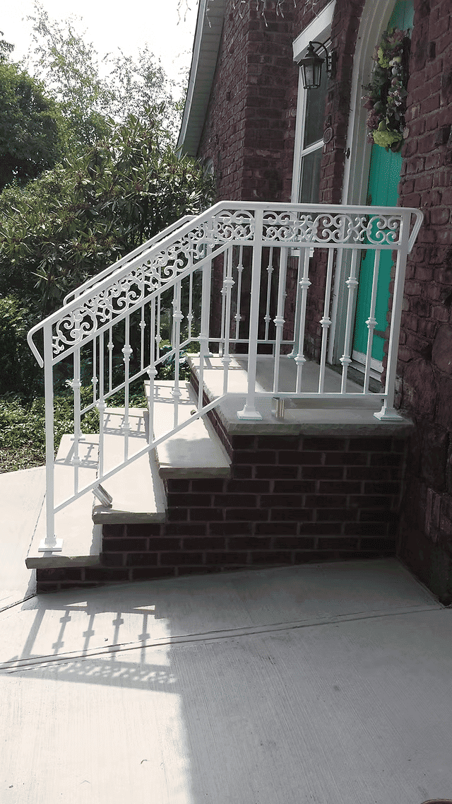 White decorative metal railing on brick steps by a green door, surrounded by greenery.