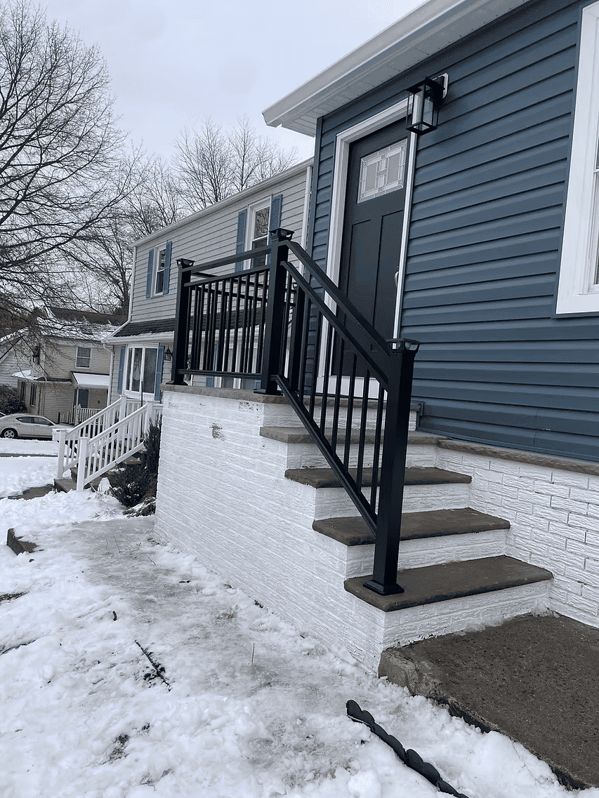 Black railing on steps of a blue house, with snow on the ground and a clear sky backdrop.