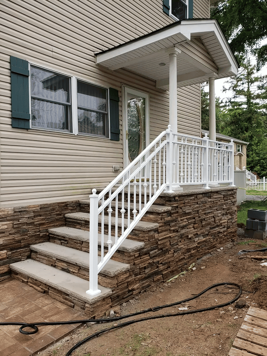 White railing and stone steps leading to a porch of a house, surrounded by greenery.