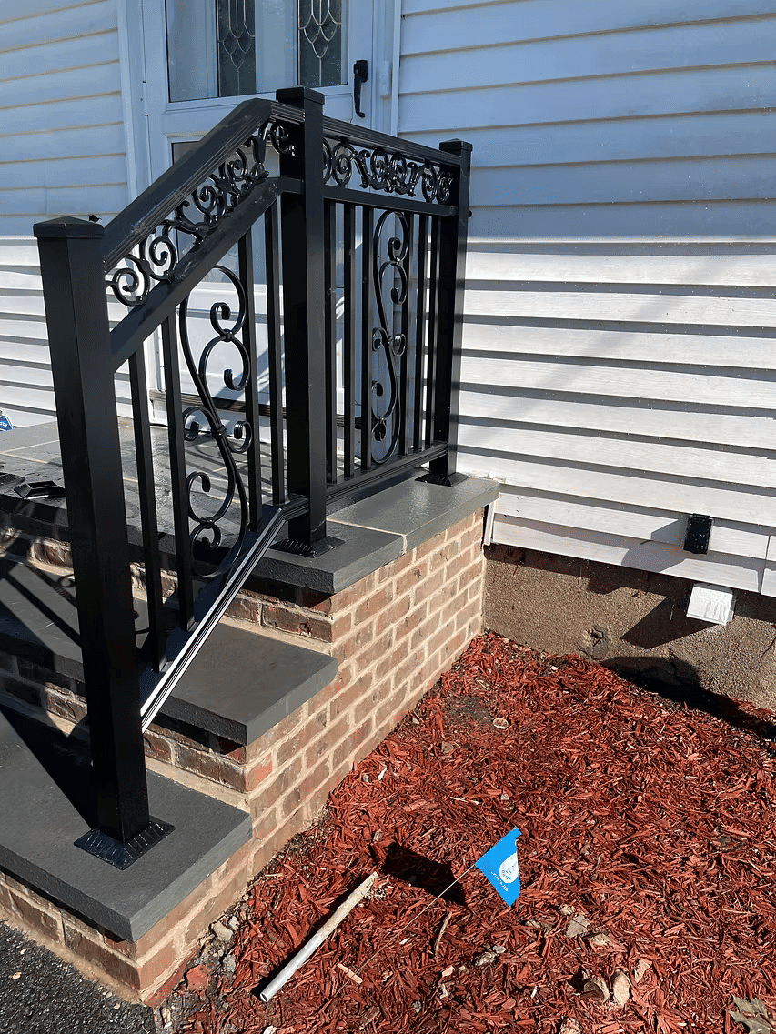 Black ornamental metal railing on stone steps of a house, with red mulch landscaping.