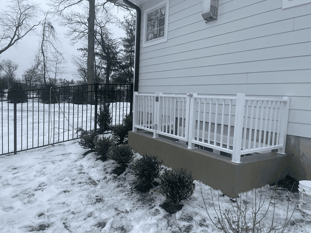 White railing on a concrete patio beside a snow-covered yard and metal fence.