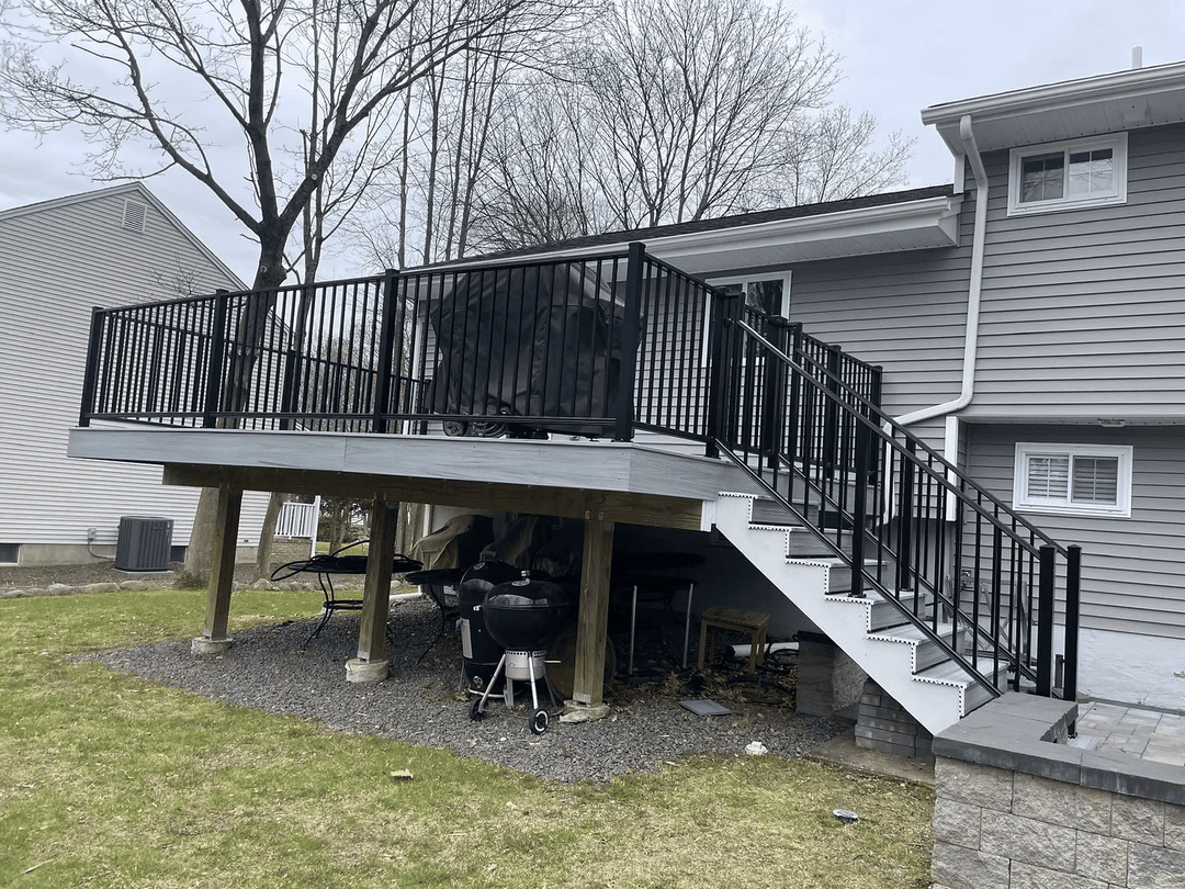 Black railing deck with stairs, surrounded by grass and trees next to a gray house.