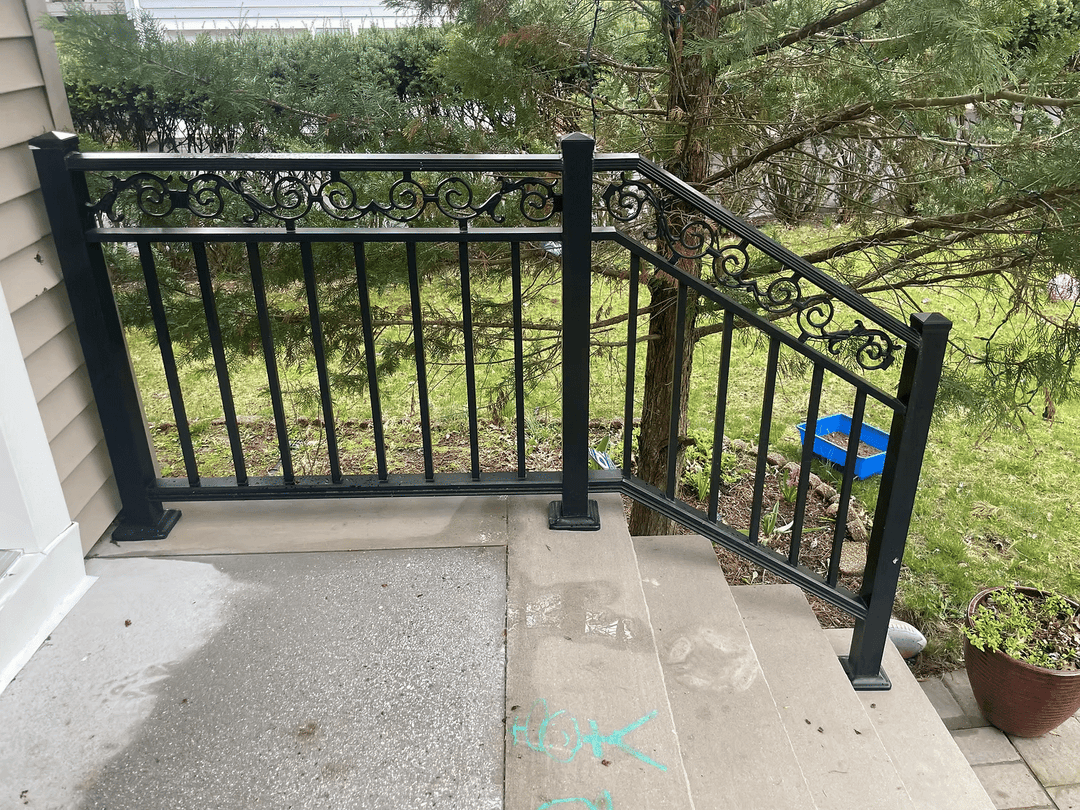 Black metal railing with ornate designs on concrete steps near a garden.